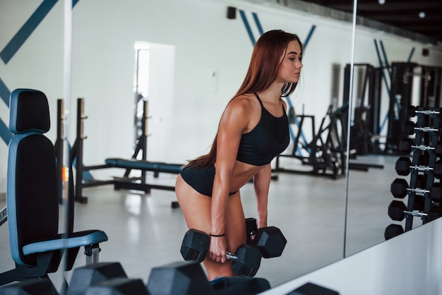 Молодая женщина фитнеса в тренажерном зале возле зеркала с гантелями в руках.