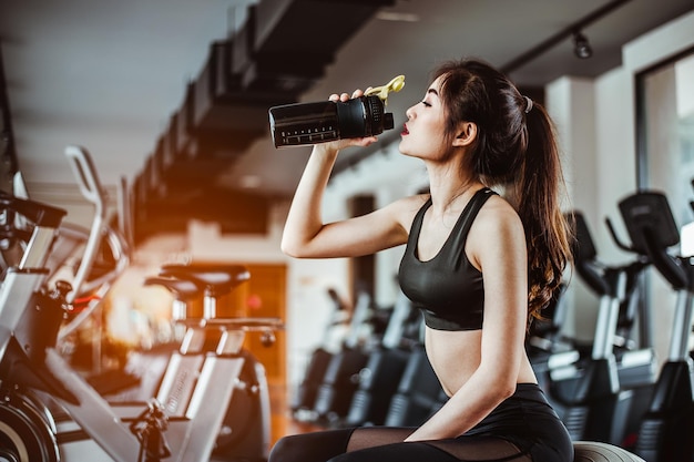 Молодая фитнес-женщина, уставшая в спортзале, пьет протеиновый коктейль, занимается концепцией фитнеса и здорового образа жизни