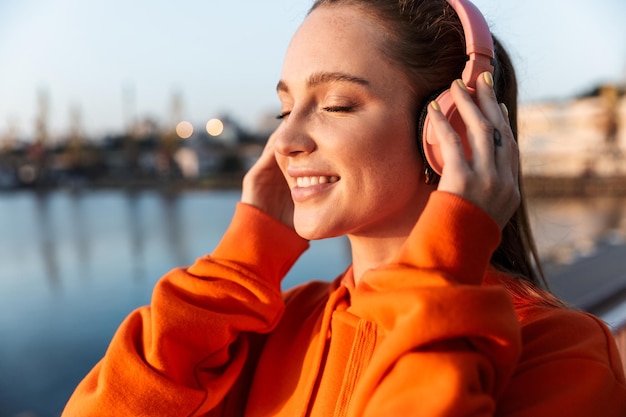 young fitness woman wearing hoodie standing at the beach, listening to music with headphones