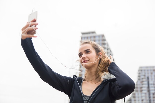 Abiti sportivi da portare della giovane donna di forma fisica che si siedono all'aperto, riposante dopo l'allenamento, prendente un selfie