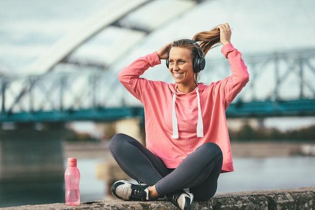 Photo young fitness woman with headphones is resting after hard training on the wall by the river with bottle of a water.