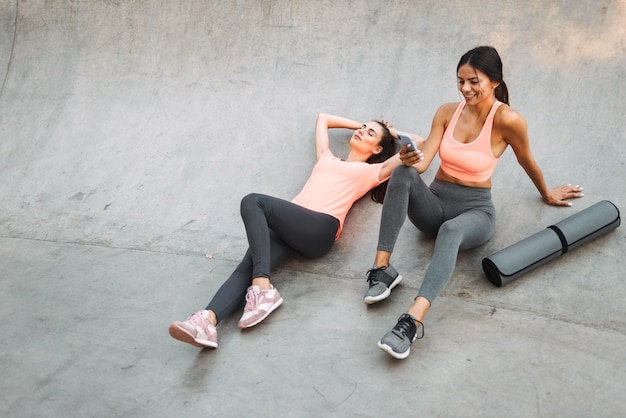 young fitness women in sportswear resting and holding cellphone while sitting on concrete sports ground