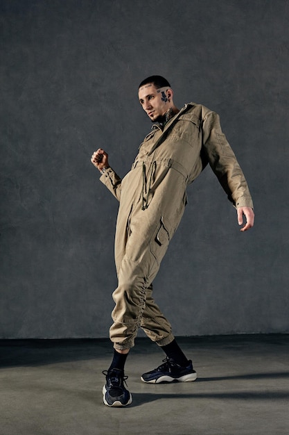 Young flexible fellow with tattooed body and face, earrings, beard. Dressed in khaki jumpsuit and black sneakers. He dancing against gray studio background. Dancehall, hip-hop. Full length, copy space