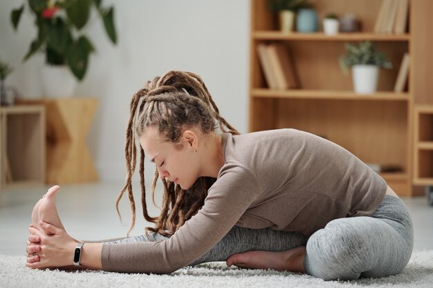 Photo young flexible woman in activewear sitting on the floor and stretching one leg forwards while bending over it