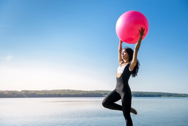 Young flexible  woman doing morning fitness exercises on pink ball near lake