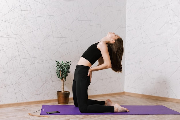 Young flexible woman practicing yoga in Supported Camel pose while balancing on mat
