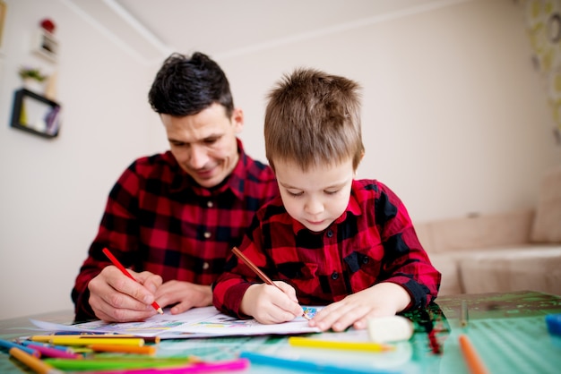 Young focused father and son in same red shirt painting with a colorful set of pencils while sitting at the table in a bright living room.