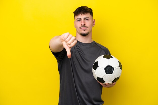 Young football player man isolated on yellow background showing thumb down with negative expression