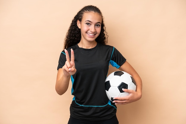 Young football player woman isolated on beige background smiling and showing victory sign