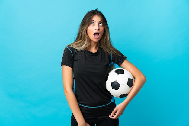Молодая женщина-футболист изолирована на синей стене, глядя вверх и с удивленным выражением лица