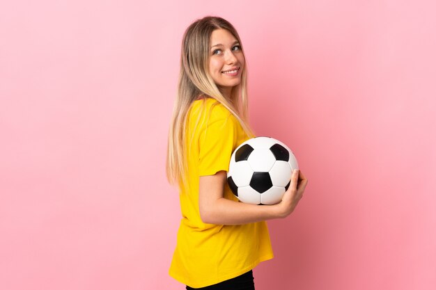 Young football player woman isolated on pink wall laughing