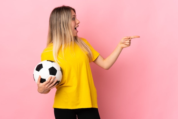 Young football player woman isolated on pink wall pointing\
finger to the side
