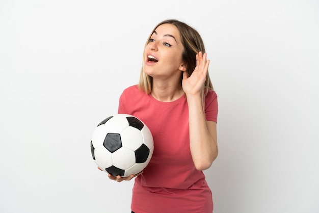 Young football player woman over isolated white wall listening to something by putting hand on the ear