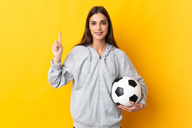 黄色の壁に孤立した若いサッカー選手の女性は、最高の兆候を示して指を持ち上げます