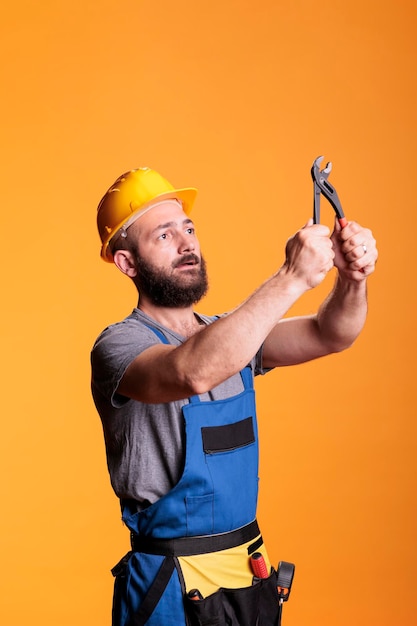 Young foreman builder looking focused at pair of pliers, using construction tools over yellow background. Male renovation expert or carpenter working with pliers for refurbishment project.