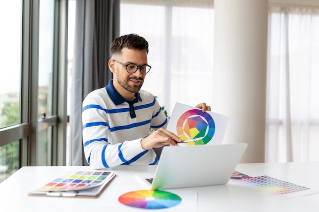 Young Freelance Graphic Designer Guy Working With Laptop And Color Swatches Sitting At Desk In Home Office Choosing Colour Gamma For New Design Project Side View With Copy Space