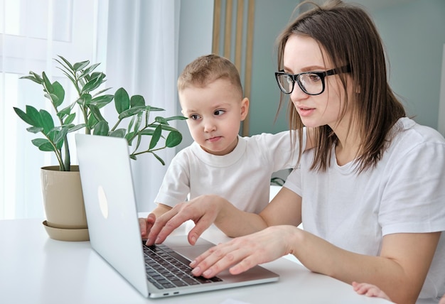 A young freelance mother works from home remotely using a laptop her son sits nearby