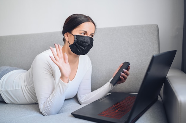 Young freelance woman in face medicine mask making a video call with laptop on grey couch during coronavirus isolation home quarantine. Covid-19 pandemic Corona virus. Online work from home concept.