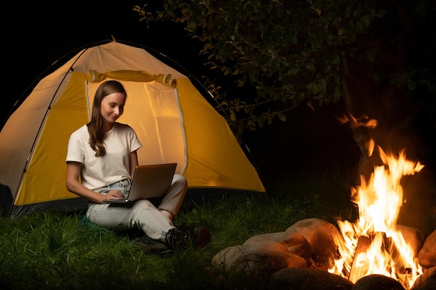 Young freelance woman working on a laptop in the evening A female tourist is sitting by a campfire and a tent