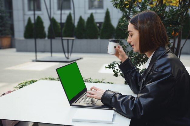 Young freelancer, business person, copywriter, beautiful brunette woman, typing text on the laptop keyboard, looking at the monitor screen with a green chromakey. Mockup, copy space to insert ads