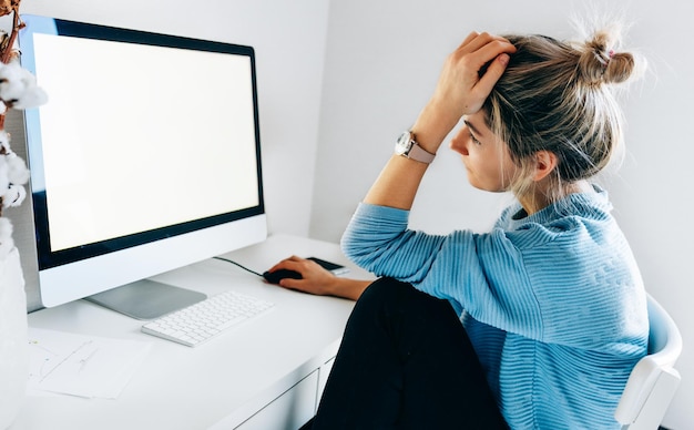 写真 オフィスでデスクトップコンピューターを使用している若いフリーランサーの実業家ブロンドの女性はあなたのテキストメッセージやコンテンツのための空白の画面でコンピューターに取り組んで自宅で屋内に座っています