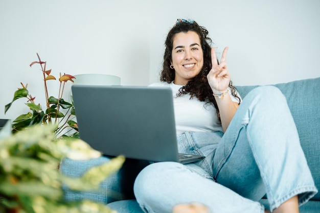 Young freelancer latina woman working on computer while sitting on the comfortable sofa at home smiling to camera doing victory sign on modern trendy clothes. Concept of remote work from home