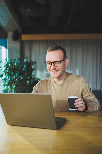 Young freelancer man in glasses sitting at a wooden table with a laptop and coffee while working on a remote project in a modern workspace