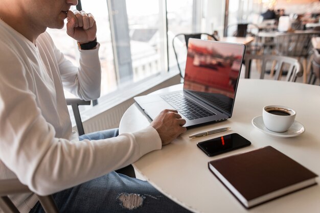 Giovane uomo libero professionista sta riposando seduto a un tavolo in un bar con un moderno laptop con una tazza di caffè con uno smartphone con un diario marrone