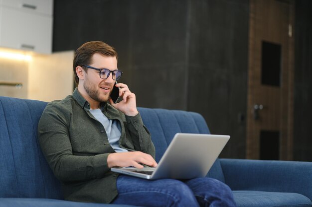 Young freelancer man in stylish glasses working at laptop computer sitting on floor at home using wireless technology for job leisure on Internet thinking touching chin