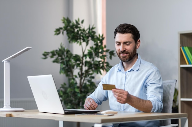 Young freelancer man working in the office at the computer at the desk Holds a credit card works with the account money makes orders