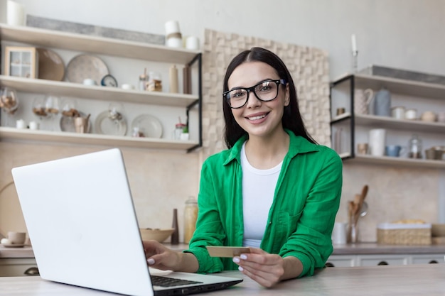 A young freelancer woman is working at home in the kitchen on a laptop and using a credit card he