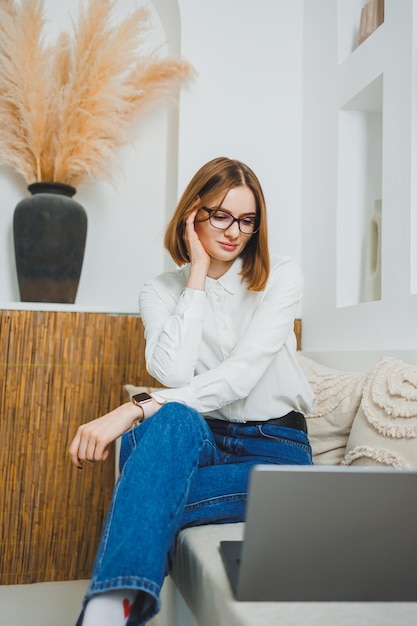 Young freelancer woman sitting on sofa and working on laptop watching movie on laptop studying blogging relaxing and chatting online