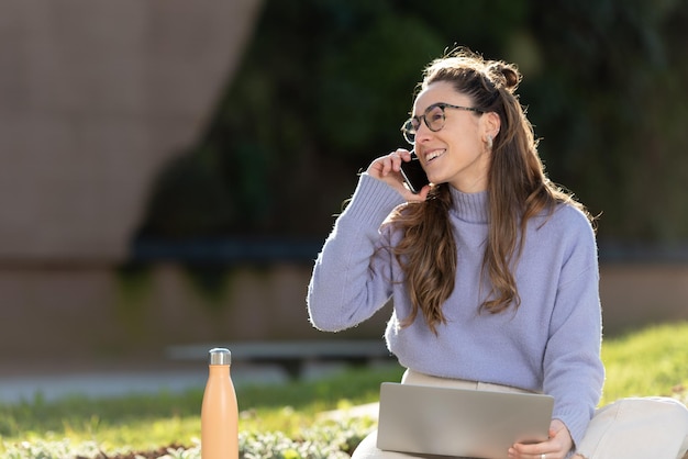 Молодая женщина-фрилансер разговаривает по телефону на улице, сидя в парке.