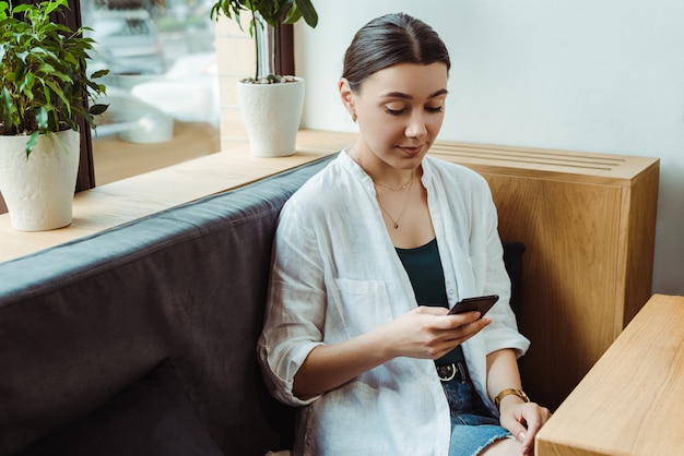 Young freelancer woman using mobile phone, planning project, researching information in modern cafe.