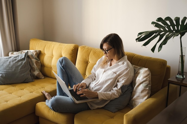 写真 屋内の黄色のソファに座って眼鏡をかけた若いフリーランサーの女性がノートパソコンと携帯電話を使って考えて作業し、距離の概念で自宅オフィスで仕事をしますコピースペース