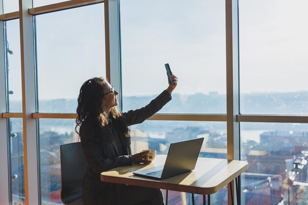 Young freelancer woman Young woman working with laptop in coworking space and talking on phone sitting in open space with cup of coffee and smiling while holding glasses