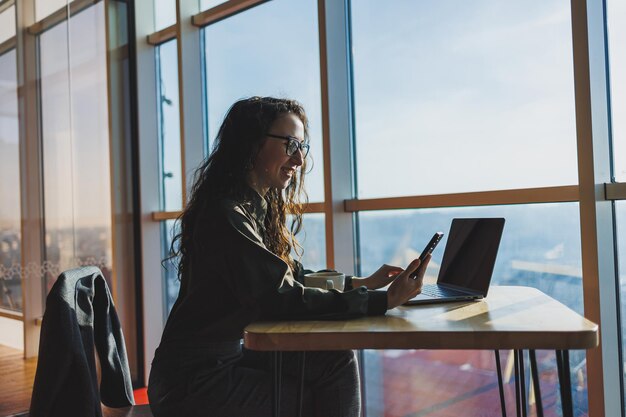 Young freelancer woman Young woman working with laptop in coworking space and talking on phone sitting in open space with cup of coffee and smiling while holding glasses