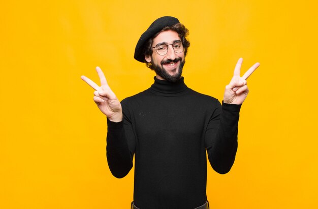 Молодой французский художник мужчина улыбается и смотрит счастливым, дружелюбным и довольным, жестом победы или мира обеими руками над оранжевой стеной