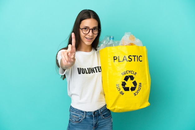 Foto giovane ragazza francese che tiene un sacco pieno di bottiglie di plastica da riciclare mostrando e alzando un dito