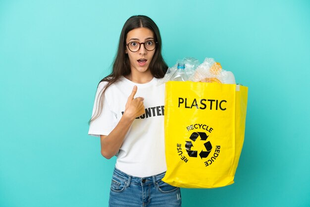 Young French girl holding a bag full of plastic bottles to recycle with surprise facial expression