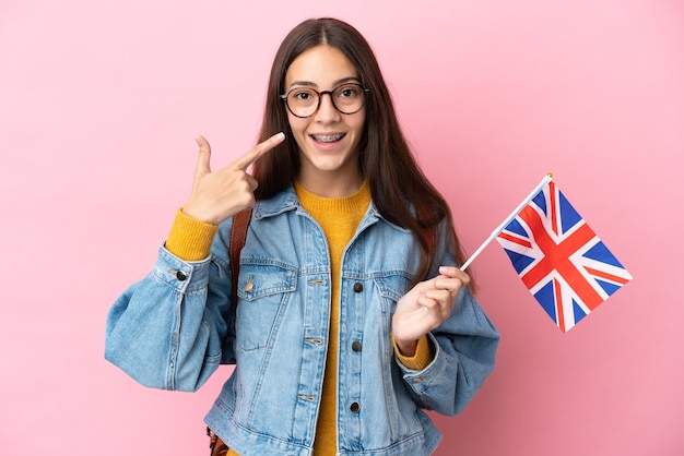 ピンクの背景に分離されたイギリスの旗を持って親指を立てるジェスチャーを与える若いフランスの女の子