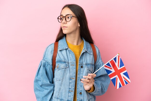 Young French girl holding an United Kingdom flag isolated on pink background looking to the side