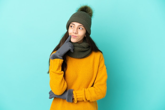 Young French girl isolated on blue background with winter hat having doubts while looking up