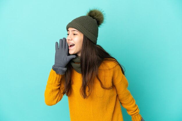 Young French girl isolated on blue background with winter hat shouting with mouth wide open to the side