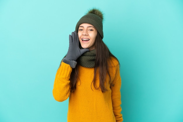 Young French girl isolated on blue background with winter hat shouting with mouth wide open