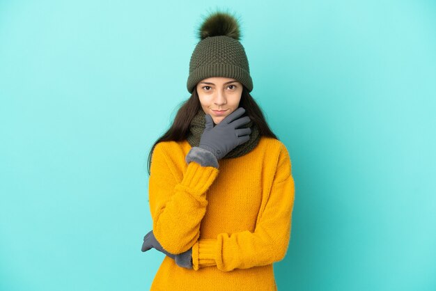 Young French girl isolated on blue background with winter hat thinking