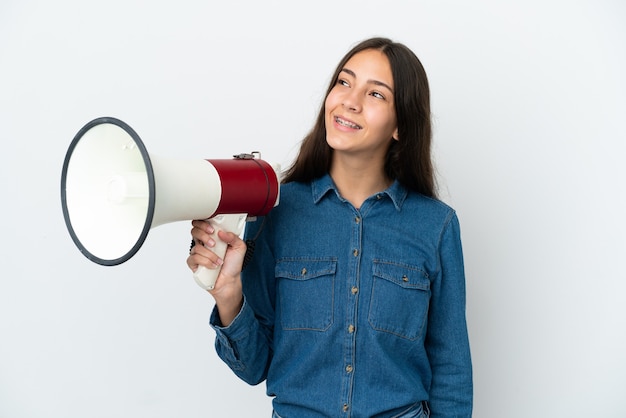 Young French girl isolated on white background holding a megaphone and looking up while smiling