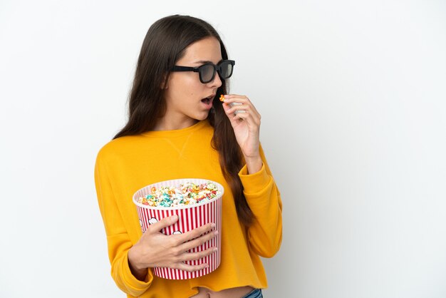 Young French girl isolated on white background with 3d glasses and holding a big bucket of popcorns while looking side