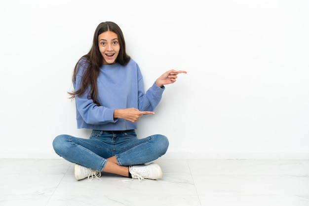 Young French girl sitting on the floor surprised and pointing side