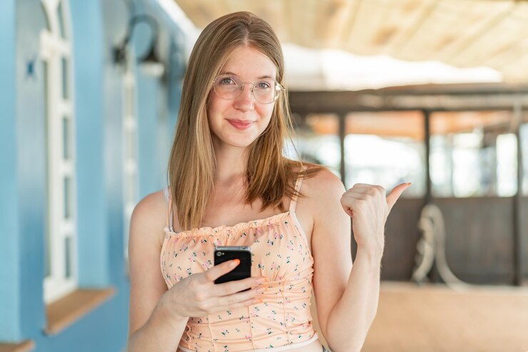 Photo young french girl with glasses at outdoors using mobile phone and pointing to the lateral